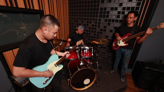 Male Asian musicians playing electric guitar, drums and bass in soundproof studio, focused on performance and teamwork, representing modern music lifestyle and creativity