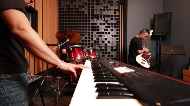 three young asian men performing in modern music studio, playing keyboard, bass guitar and drum kit together, creating a focused and professional rehearsal atmosphere