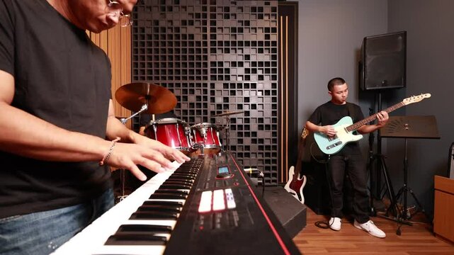 Tight shot of Asian male playing digital keyboard, guitarist in background, red drum set and studio equipment visible, musical synergy