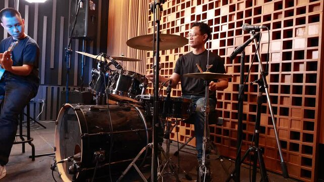 two young asian male musicians playing electric guitar and drums in professional soundproof studio, deeply focused on live performance session surrounded by recording equipment
