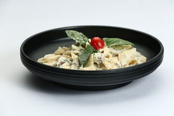 Creamy pasta in black bowl: penne with white sauce, herbs, cheese, and a tomato on a white background