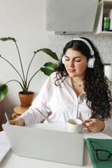 Vertical image of intelligent concentrated young Caucasian female using education platform studying online on laptop in headphones, making notes while listening to lecture or watching video tutorial