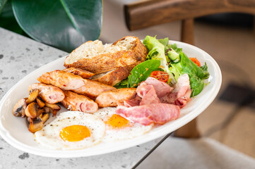 A generous breakfast plate features fried eggs, sausages, crispy bacon, sautéed mushrooms, and fresh salad, served with toasted bread. The vibrant presentation invites a satisfying meal.