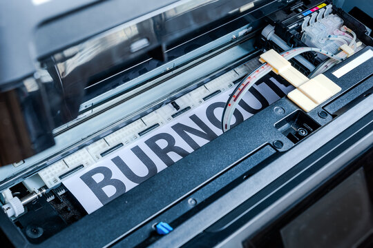 Photo from inside a printer as it prints the word burnout on white paper &mdash; a metaphor for mental exhaustion and the need for regular human maintenance, like machines.