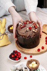 Woman in white shirt hold Easter Easter cake,Easter bread with chocolate glaze decorated with dried cherries and walnuts on a light background,tall candles, flowers, Easter bunny nearby.Easter concept