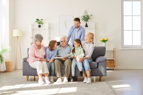Big family of different generations gathering on sofa around grandfather, looking through photo album. Parents, children and grandparents enjoying leisure time and good memories together at home.