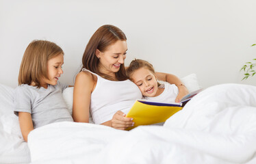 Mother and children relaxing together in bed at home, reading a book in the cozy bedroom. The family enjoys a peaceful moment of leisure, sharing time and stories in a comfortable setting.