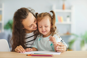 Portrait of happy family sitting together at desk with smiling mother hugging her preschool daughter who drawing picture with colorful pencils. Loving mom developing creativity of her cute child.