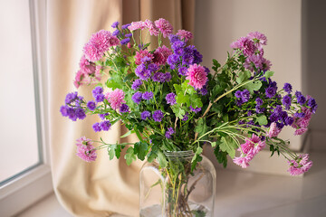 A bouquet of lilac and pink chrysanthemums in a glass jar by the window.