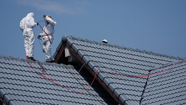  house painter is standing on a roof while using an electric power spray painter on roof .