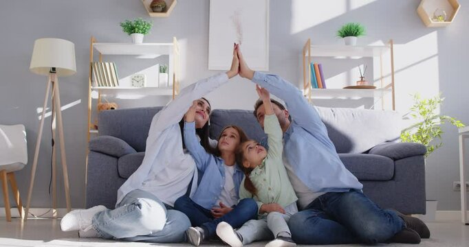 Loving young parents sitting on floor at home, putting hands in stack and forming roof shape with arms above their smiling son and daughter. Family safety, protection, love and secure future together.