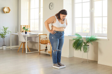 Overweight young woman standing on scales at home, looking down and checking progress of weight loss. Fat female measuring weight, tracking fitness and weight goals during weight loss and slimming.