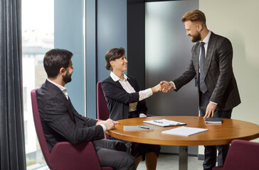 Corporate colleagues coworkers sitting at round table and making hand shake gesture after negotiations, meeting in office, look gladfully at each other, two men and woman gathering at briefing
