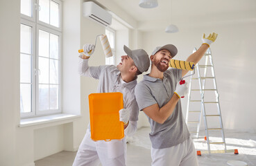 Team of happy professional house painters holding paint rollers as if microphones and singing...