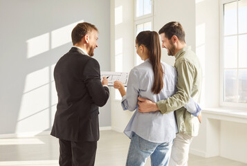 Fototapeta premium Back view of real estate agent in suit presenting apartment layout to happy young couple in bright empty room. Smiling man and woman hugging each other, discussing future home purchase with realtor.