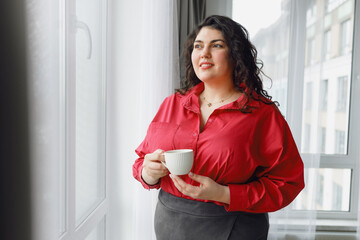 Side view of plus size businesswoman in pink shirt having coffee at home, admiring cityscape...