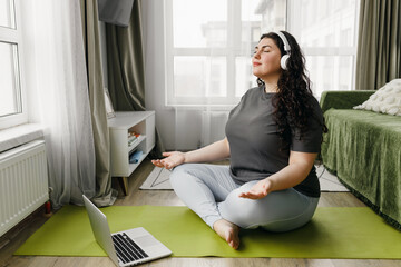 Plus size woman taking care of her body and mind, sitting on floor on mat in lotus posture with...