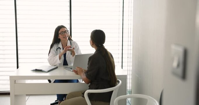 Young female doctor dressed in white coat talking with patient, provide medical advice, treatment plan of heath recommendation seated at desk in examination room. Professional healthcare consultation