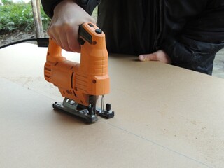 hand presses orange jigsaw and moves along marking line, cutting sheet of pressed wood into pieces,...