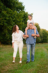 Family walking in park. Dad is carrying baby girl on neck, mom is walking next to him, smiling happily. Family is having fun together, walking along path outside. Happy family, parental care concept.