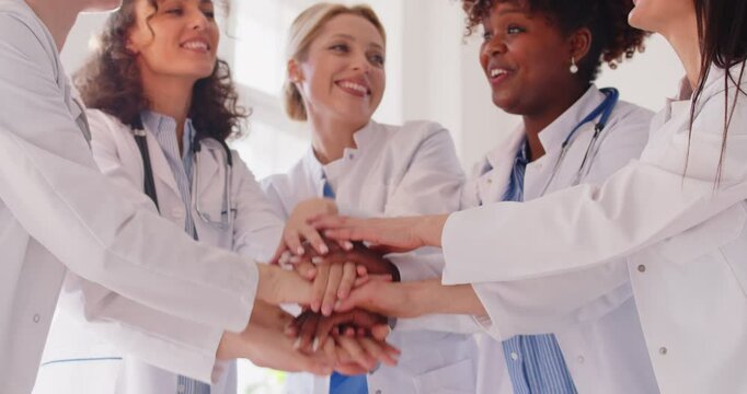 Group of cheerful female doctors in white coats stacking hands together and clapping in medical office. Smiling healthcare professionals celebrating success, showing teamwork, unity and motivation.