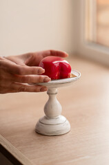 Close-up of female hands placing glossy red heart-shaped dessert on vintage white stand near window, soft natural light, cozy and elegant scene