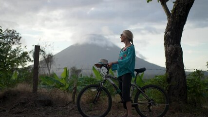 A senior woman smiles while biking near Concepcion volcano on Ometepe Island, Nicaragua. Lush greenery surrounds her as clouds gather overhead. - Powered by Adobe