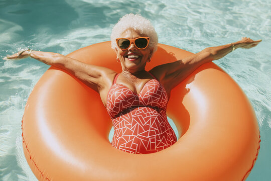 Senior woman with white hair wearing sunglasses and red swimsuit floats on orange inflatable ring in turquoise pool, enjoying summer relaxation