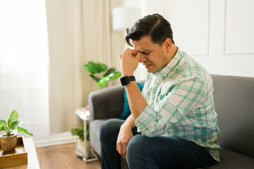 Stressed man suffering from a migraine and touching his forehead while sitting on the sofa in the...