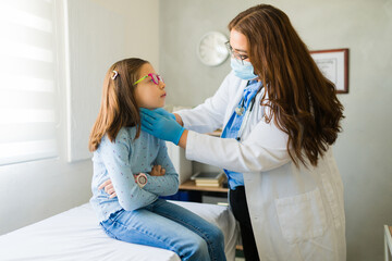 Female doctor wearing face mask and gloves examining throat of sick girl sitting on examination...
