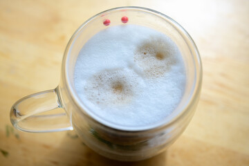 Foamy Cappuccino in Clear Glass Cup on Table
Top view of a cappuccino with thick foam served in a transparent glass mug, resting on a wooden table in natural light.
