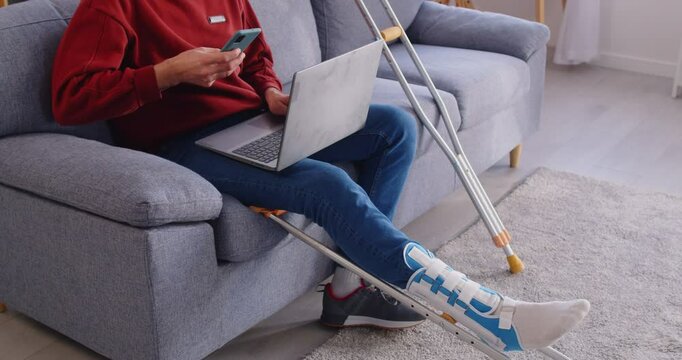Broken leg trauma, foot injury man sitting on sofa, working with laptop online, crutches with bandaged at home, patient rehabilitation, person not be able to walk, severe fracture, protective cover