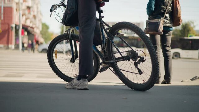 Cyclist with backpack walking next to bicycle in urban setting. Street view with motion blur, focus on shoes, bike wheels, and surroundings. City scene with crosswalk, sunny weather, casual style