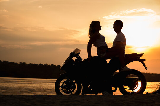 young beautiful pair of lovers with nice figures and silhouettes, posing in the sunset on the ocean sitting on a retro motorcycle caferacer, outdoor portrait,