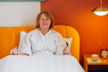 An elderly woman in a white coat is sitting in bed and meditating. A mature lady puts her fingers together in a meditation pose, closes her eyes and relaxes before going to bed.