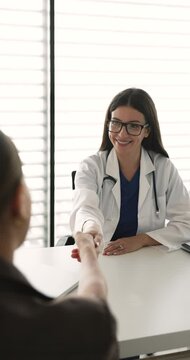 Smiling female doctor dressed in white coat sitting at desk in examination room, handshaking with woman patient express friendly attitude, start or finalize successfully appointment in clinic office