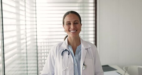 Head shot of smiling woman doctor stands confidently in modern medical office, wear white lab coat and stethoscope around neck, posing for professional portrait, exuding professionalism and competence