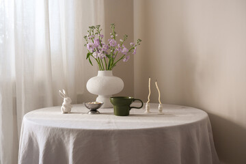 A beautifully easter serene table arrangement featuring vibrant flowers, elegant green cup of coffee, ceramic rabbit, and minimalistic candles on round table.