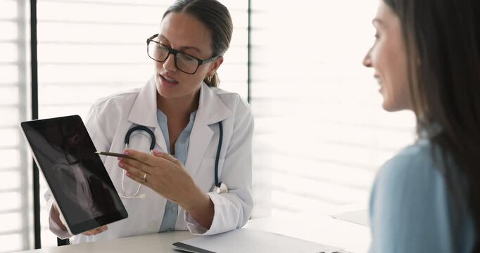 Traumatologist in white coat holds tablet and pointing at X-ray image displayed on screen, explaining xray results, describe diagnosis, injury, or treatment plan to woman patient. Healthcare services