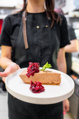 A server presents a slice of creamy chocolate cheesecake garnished with vibrant berry compote and a sprig of mint. The elegant dessert showcases a blend of textures and flavors in a stylish setting.