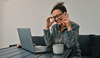 Woman Holding Phone and Smiling While Working on Laptop at Home