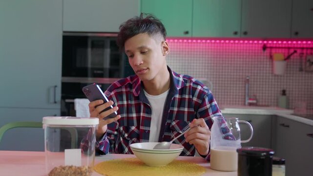 featuring focused mixed-race man in plaid shirt scrolling phone during breakfast. checking emails, chatting work messenger with cereal in cozy stylish kitchen, calm lifestyle morning moments at home