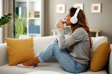 Young woman relaxing on sofa drinking coffee and listening music with wireless headphones