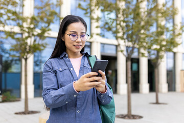 A young asian woman with glasses looking at her phone in front of a building. She is wearing a blue...