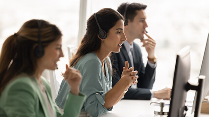 Serious Latin telephone assistant woman sitting at workplace with contact center colleagues,...