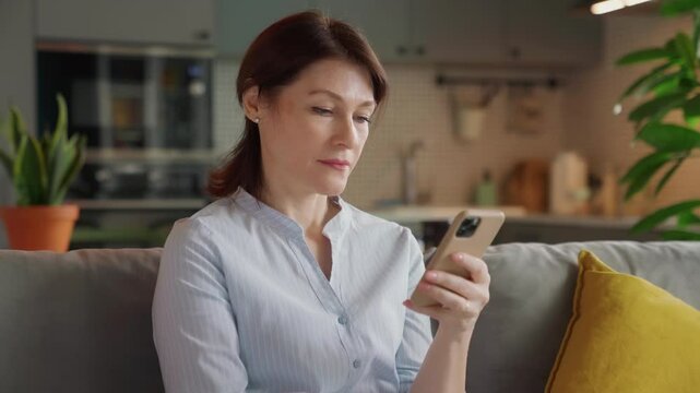 excited middle aged woman saying wow while looking at smartphone screen. happy moment of surprise and joy on cozy couch in stylish modern kitchen, natural light, real-life reactions at home