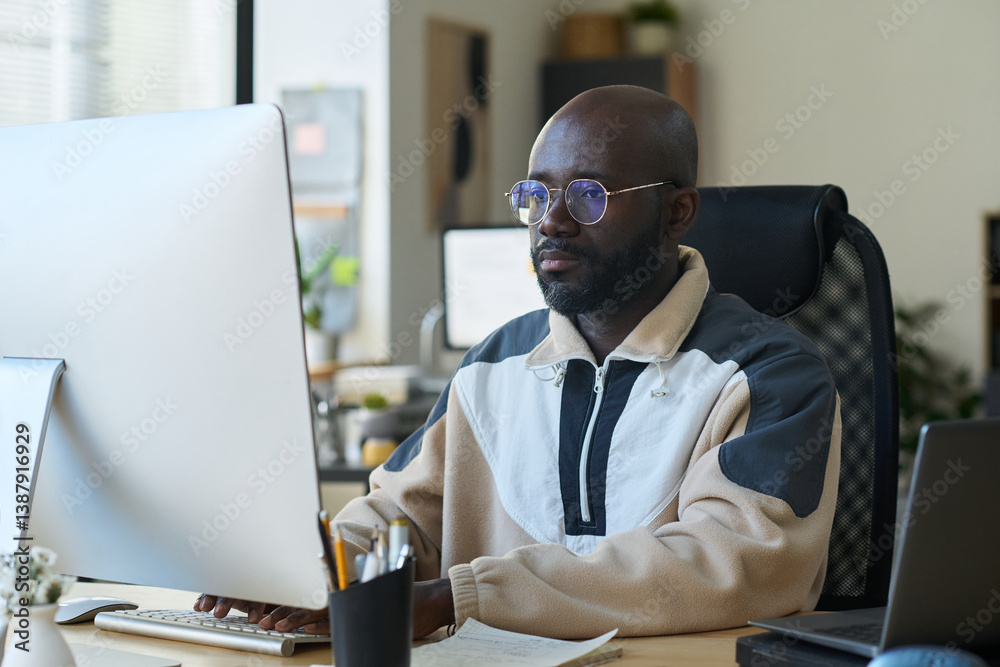 Aufkleber young serious male designer in eyeglasses and casual attire looking at computer screen and pressing 