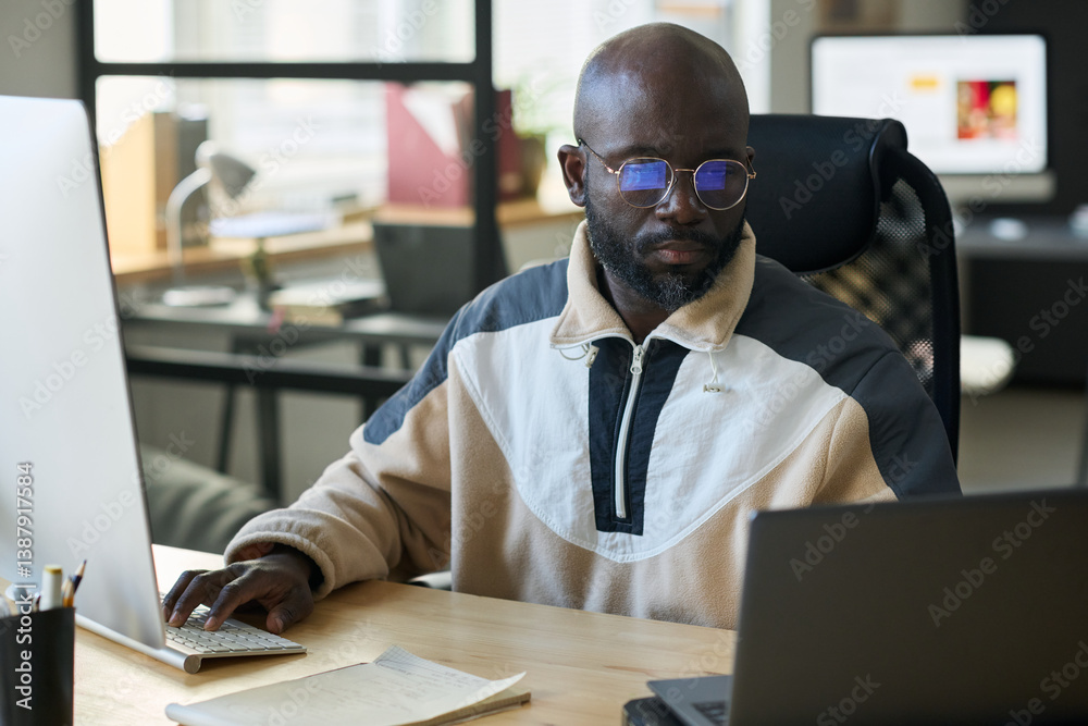 Poster Young African American man looking at laptop screen and touching keypad of desktop computer while networking in office