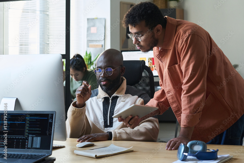 Aufkleber serious young man with tablet bending over desk while standing next to colleague and both looking at