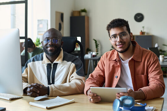 Two happy young intercultural male programmers looking at camera while using desktop computer and tablet in coworking space